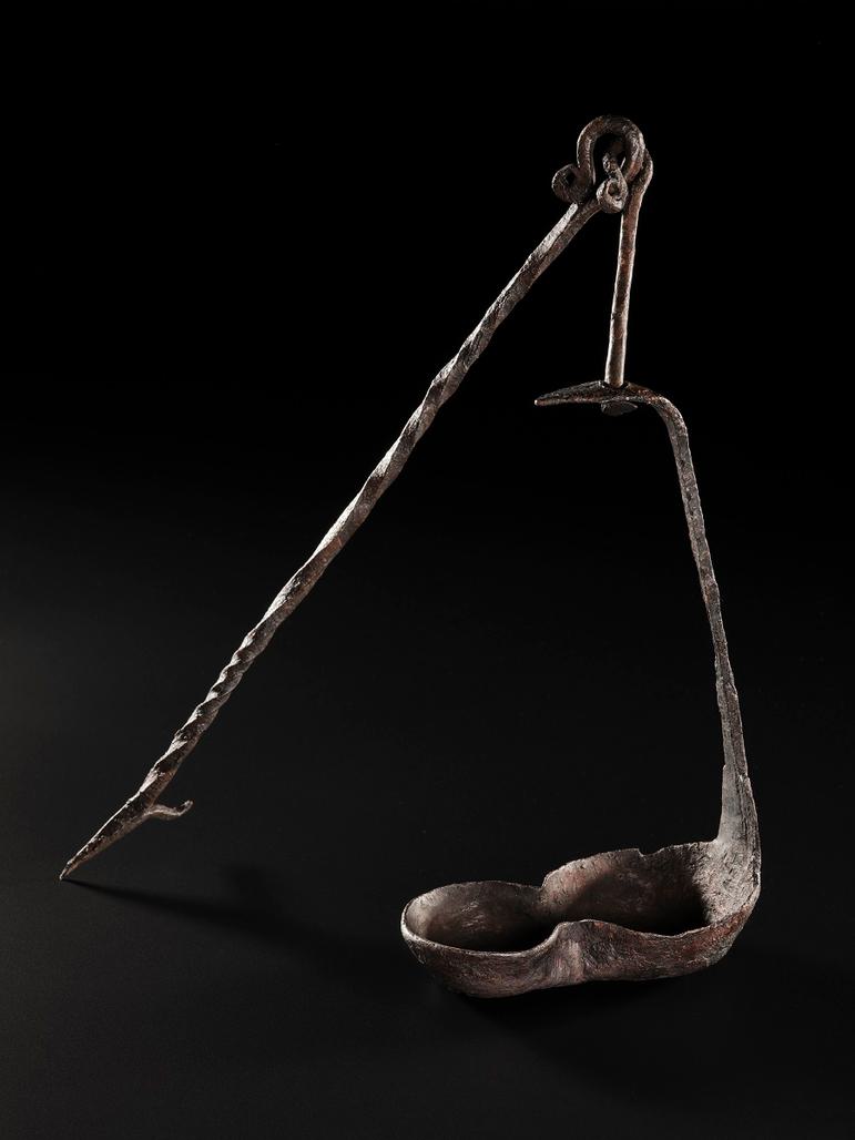 Dark brown lamp with figure-eight basin, long neck pointing up terminating at a loop, and a spear-like appendage pointing diagonally down to the left.
