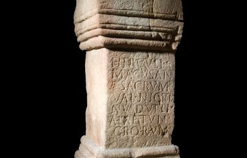 A small top and bottom-heavy dusty yellow pillar with faint Latin script written in neat lines in the thinner, middle section.