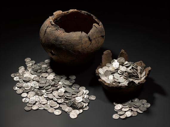 On left, a large, empty dark brown pot missing its lid with a pile of silver coins in front of it. On right, the basin of another container filled with silver coins.
