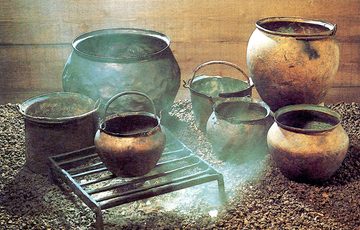 Five simple cooking pots inside a wooden building, one of which is over a grill with smouldering rocks under it.