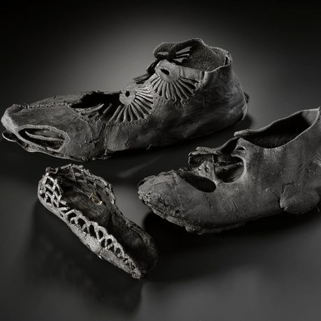 Set of three blackened leather shoes on a dark surface. A large, decorated men's shoe, a medium-size women's shoe, and in front a small child's shoe of mostly just the sole.