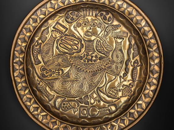 A round gold object with intricate designs engraved including a stylised person at the centre
