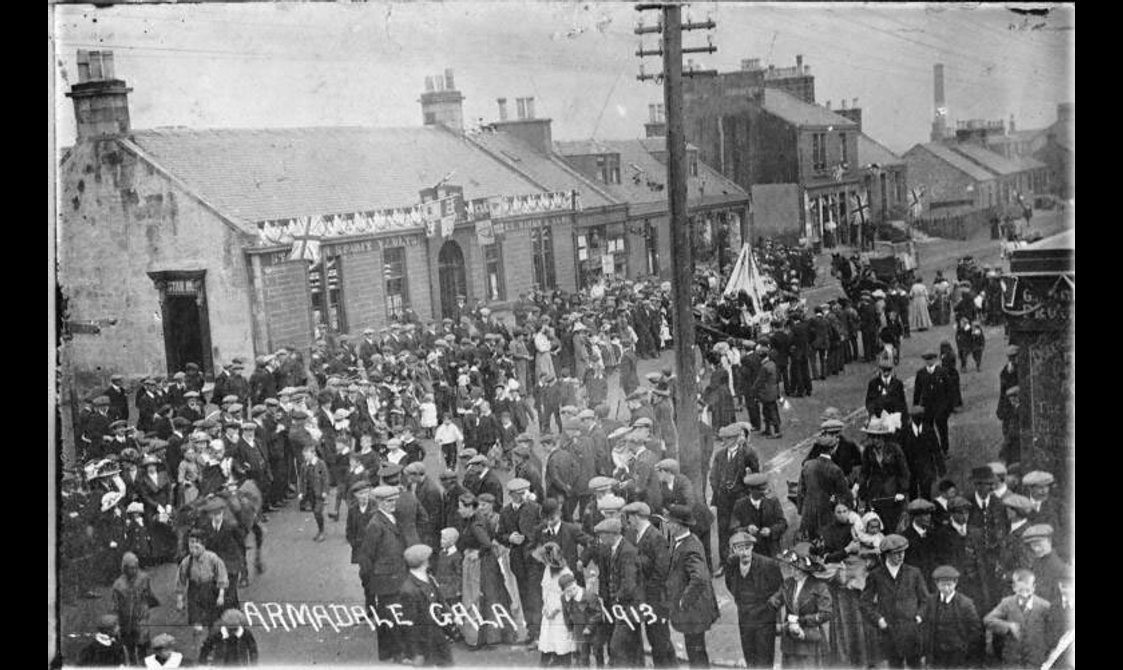 Black and white photograph. Crowds line the street to watch the procession. Buildings along the procession route are flying flags and bunting for the occasion