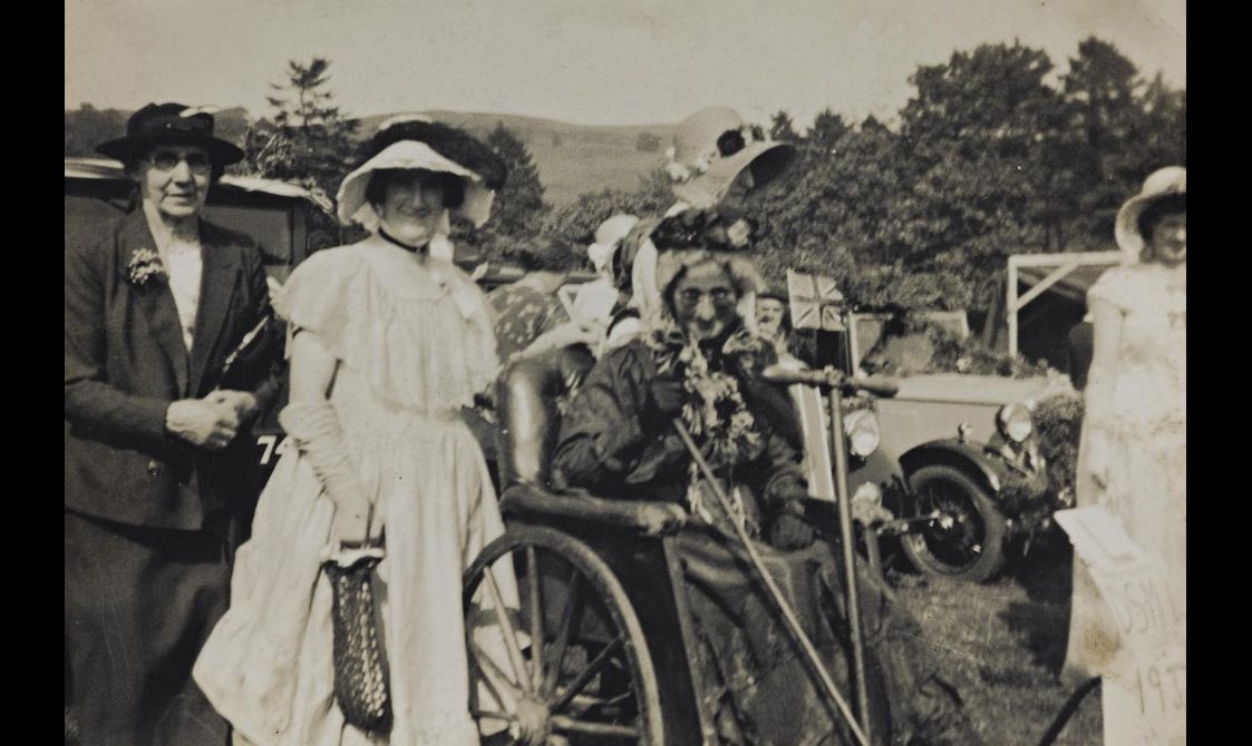 A group of five women wearing dresses and hats. One of the women is in a wheelchair.
