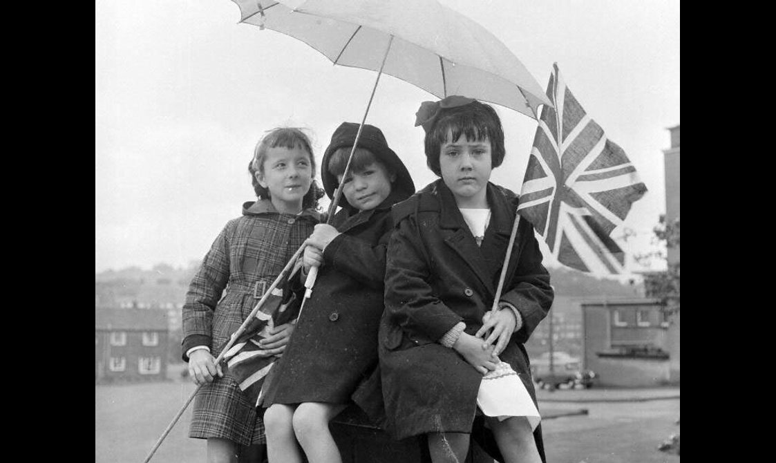 Black and white photograph of three children sitting under an umbrella in woolen coats