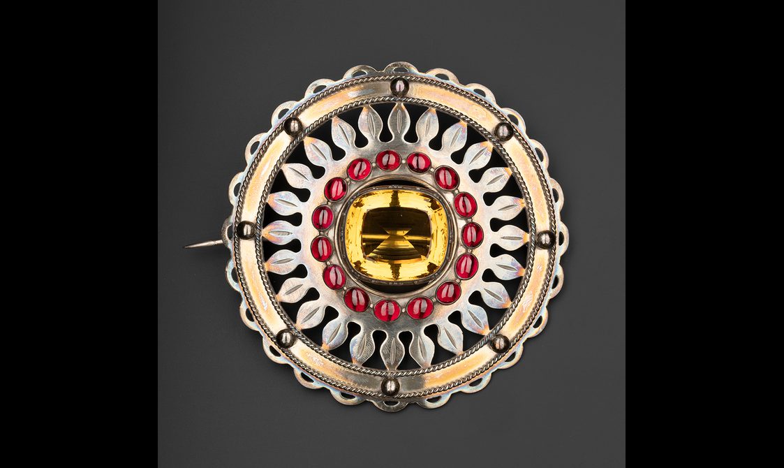 A round silver brooch with a yellow cairngorm in the centre and a ring of sixteen red stones round it.