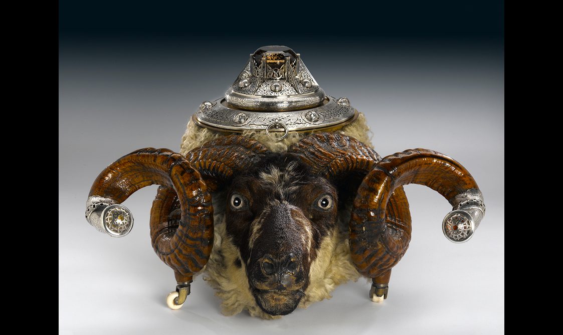 A snuff box made from a ram's head. A silver lid adorned with an orange cairngorm sits on the top of the head. Its curls horned feature silver mounts.
