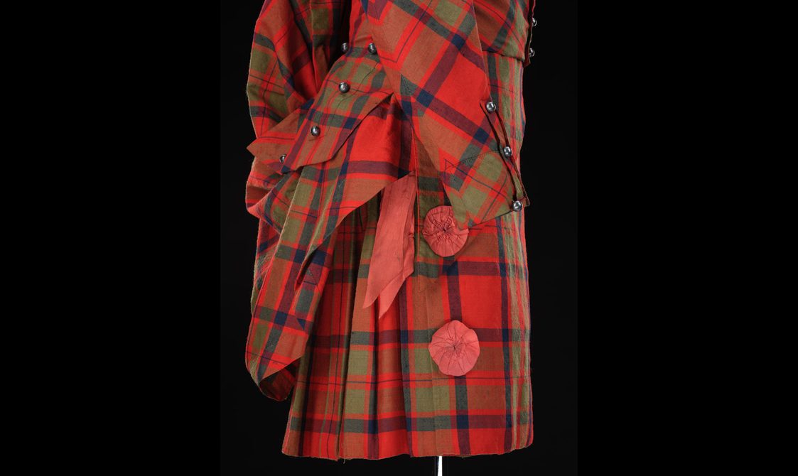 detail of a mannequin wearing a red tartan kilt with a matching jacket with layered coattails and rosette decoration.