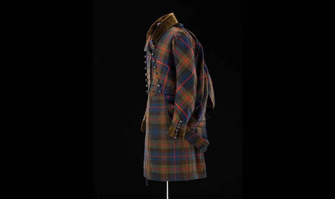 Side view of a mannequin wearing a navy, green, and red tartan kilt with a matching jacket