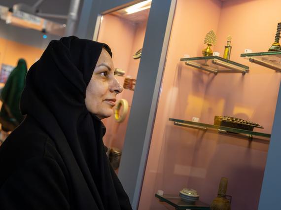 A profile of a woman wearing a black hijab (material covering over her head and shoulders) intently looks at golden and other small objects from west and central Asia.