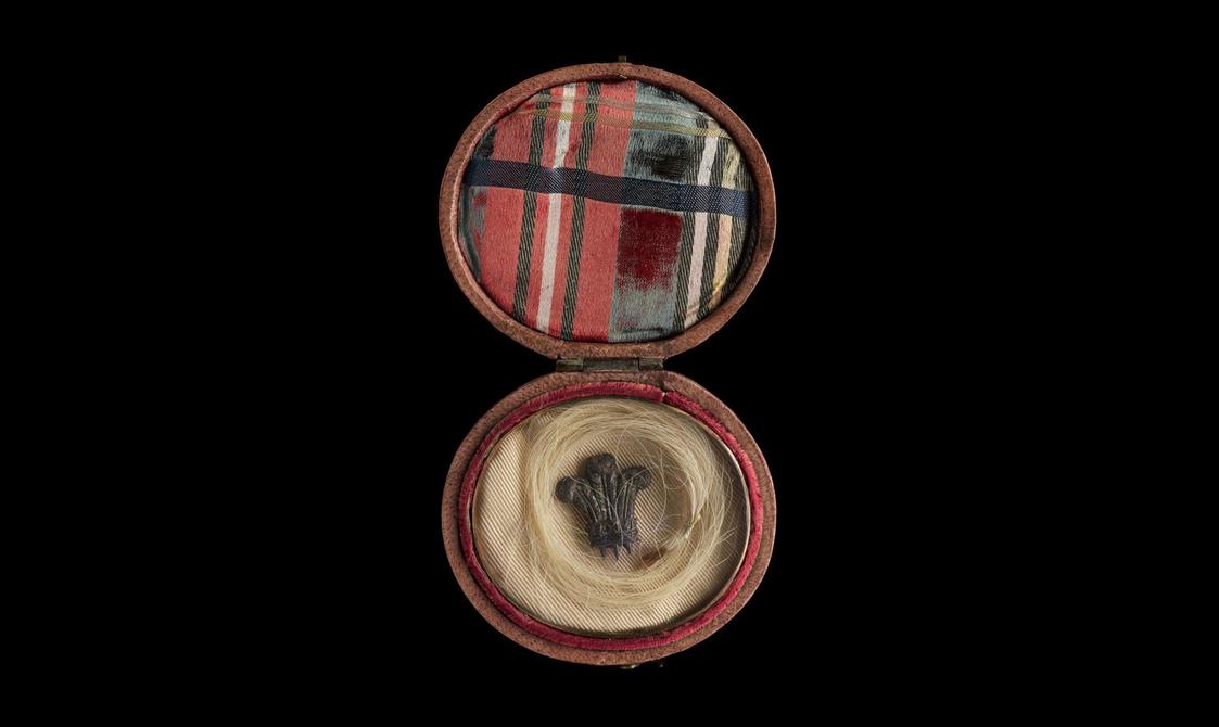 An open round box with a tartan fabric attached to the inner lid of the box, and a lock of blonde hair inside. A small metal item in a shape of three feathers placed in the center of the lock. Set against a black background.