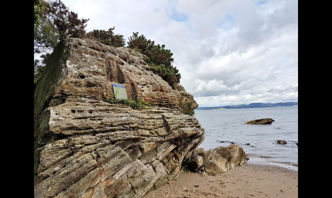 15ft-tall grey-yellow rocky outcrop next to the water, with a small figure carved on its upper face within a shrine-like recess.