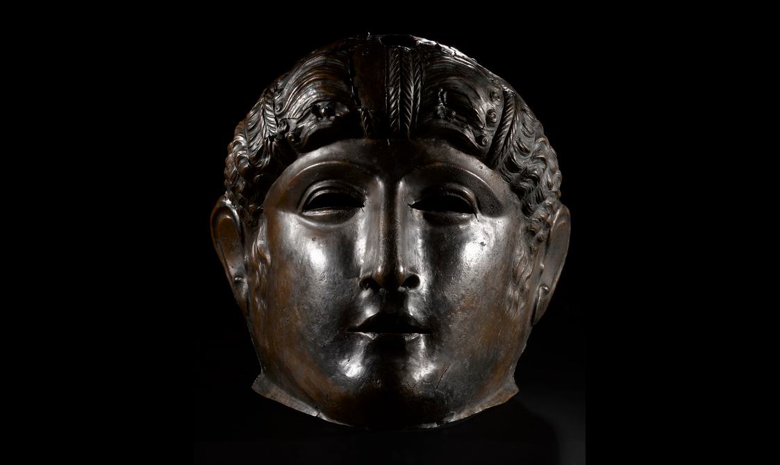 Frontal view of a dark bronze mask, as though speaking to someone standing close. Broad cheeks converge on a small mouth, strong nose and stoic eyes.