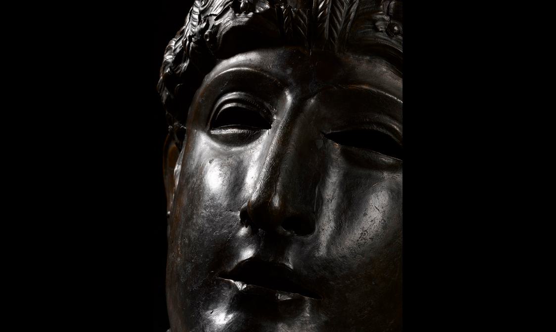 Close-up of a dark, bronze mask, lingering on the eyes and mouth which convey a stoic if somewhat sad expression.