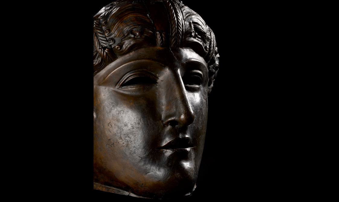 Profile view of a dark bronze mask with full cheeks, a strong nose, stylised wavy hair and air of dignity.