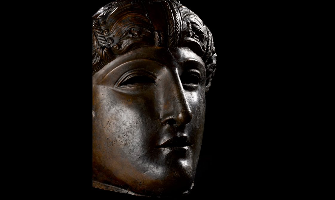 Profile view of a dark bronze mask with full cheeks, a strong nose, stylised wavy hair and air of dignity.
