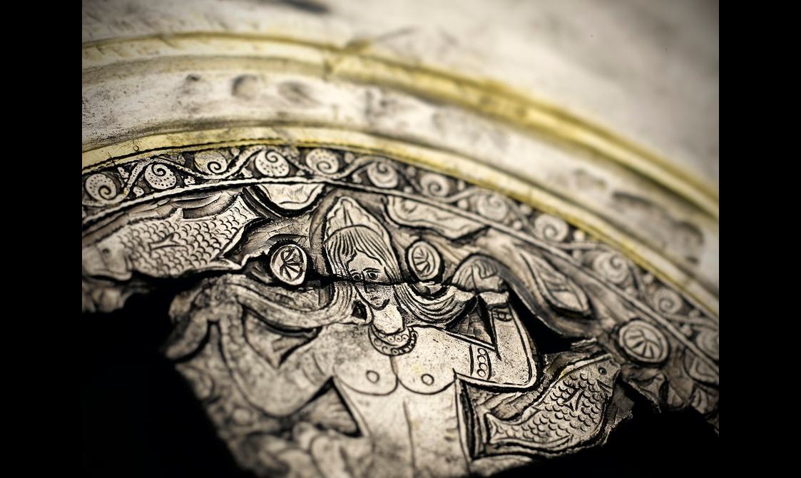 Extreme close-up up a silver dish detail showing a male god rising out of stylised waves underneath an arch of geometric designs.