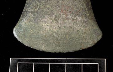 The edge of a weathered bronze axehead with white ruler measurements against it