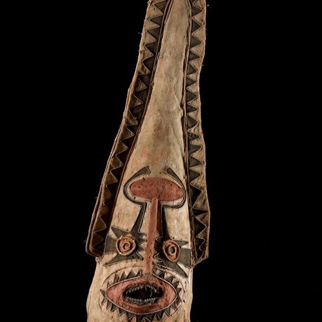 A ceremonial mask with a long tapered head. It is light brown with black and red details around the mouth, eyes, and edging.
