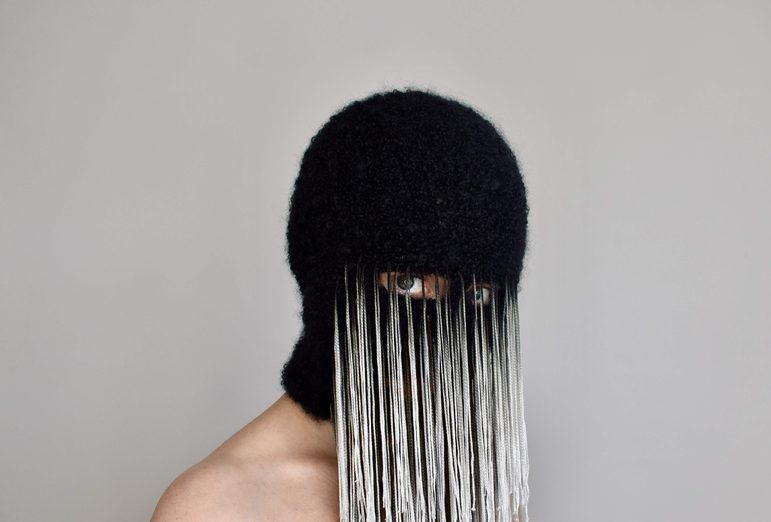 A person wears a black mask that covers their face except for their eyes. There are long white and gret threads attached from the eye openings.