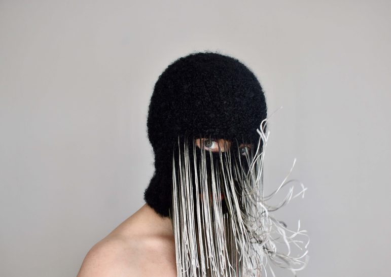 A person wears a black mask with only their eyes visible. There are long white and grey threads attached to the mask that move with the person's breath.