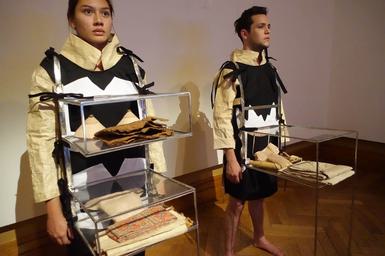 Two people stand beside each in a gallery other wearing brown tunics and clear display cabinets attached to their chests.