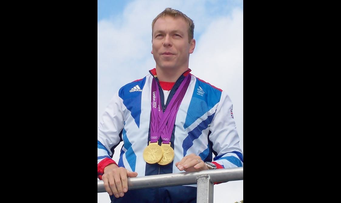 A man with short blonde hair wears a sports jacket with two Olympic gold medals around his neck.