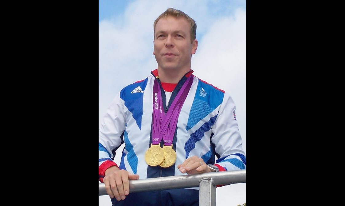A man with short blonde hair wears a sports jacket with two Olympic gold medals around his neck.