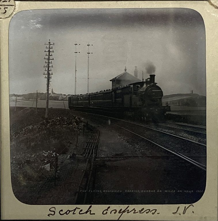 A black and white square slide of a steam train moving on a train track through the countryside.