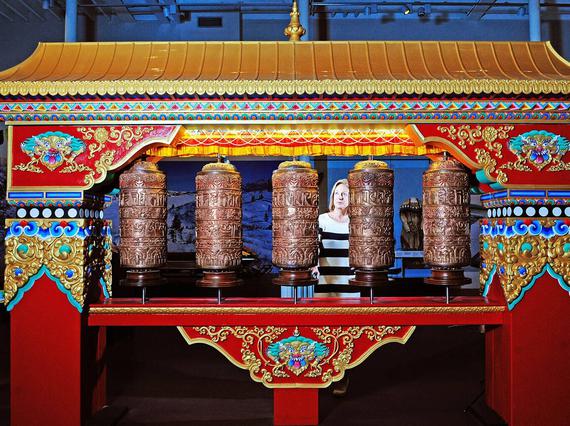 A woman stands behind a large Tibetan structure that is red with a gold roof and decorated pillars. It has five brown spinning prayer wheels at its centre.