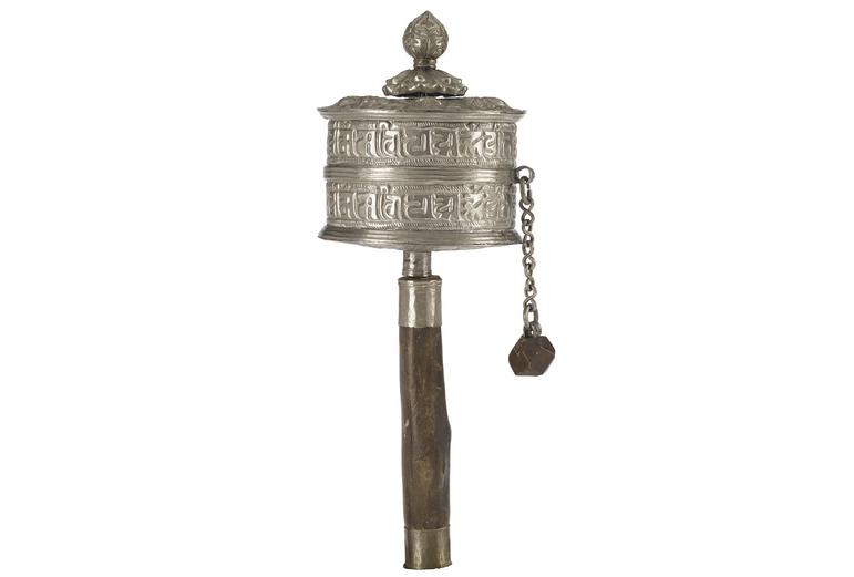 A round silver lidded case with Tibetan script embossed on its surface attached to a wooden handle. There is a small chain with a piece of wood at its end attached to the silver case.