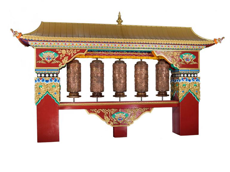 A house like frame with a gold roof and red pillars and decorated surfaces. There are five brown spinning prayer wheels inside.
