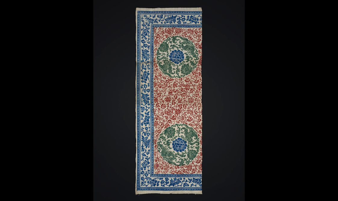 Sample of printed cotton with an ornate red ink design featuring a circular green and blue feature with a blue border