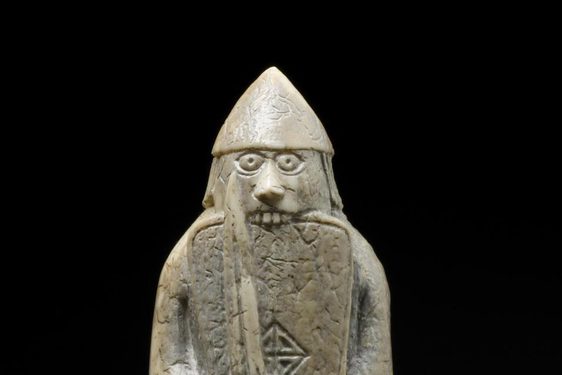 An ivory chess pieces of a figure wearing long robes and a helmet while holding a sword and shield