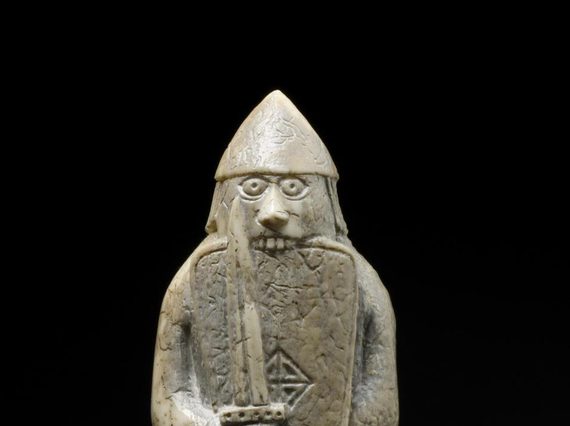 An ivory chess pieces of a figure wearing long robes and a helmet while holding a sword and shield