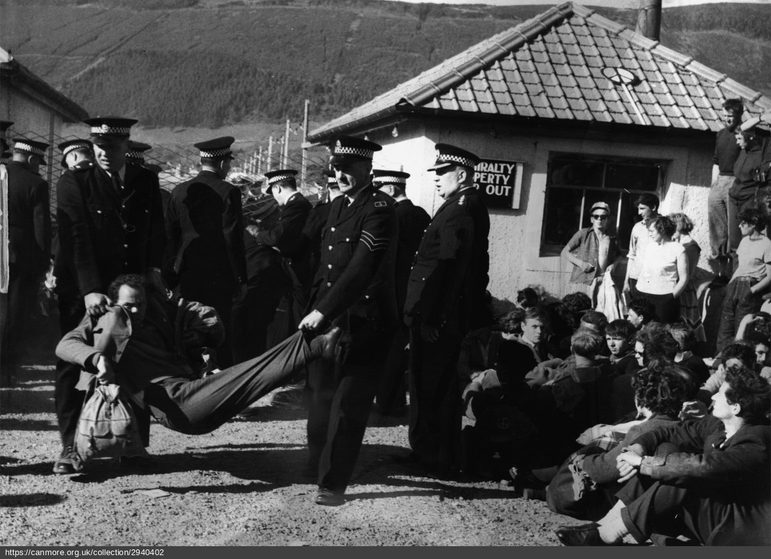 A black and white photograph showing policemen carrying a protester in front of a small property. Other protesters are seated on the ground, looking on.