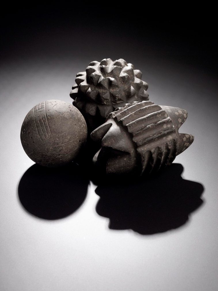Three grey stone balls, one shaped as a circle with linear decorations, one carved with protruding triangles, and another carved out of triangles to the ends and linear decorations.