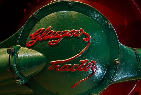 A colour photograph of a close up of a green tractor plate with the Glasgow tractor red painted logo. The logo is in a hand-lettering style.