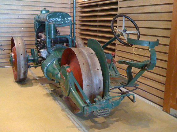 A colour photograph of a green Glasgow Tractor with 2 metal wheels at the front and 1 wheel at the back. The wheels are painted red on the inside. There is an engine above the 2 front wheels and a steering wheel and seat at the back wheel.