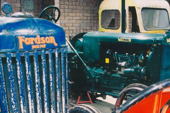 A colour photograph of 4 different colour tractors: A blue Fordson, a green Wallis and a partial view of another yellow tractor in the background and a bright orange tractor on the right