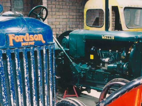 A colour photograph of 4 different colour tractors: A blue Fordson, a green Wallis and a partial view of another yellow tractor in the background and a bright orange tractor on the right