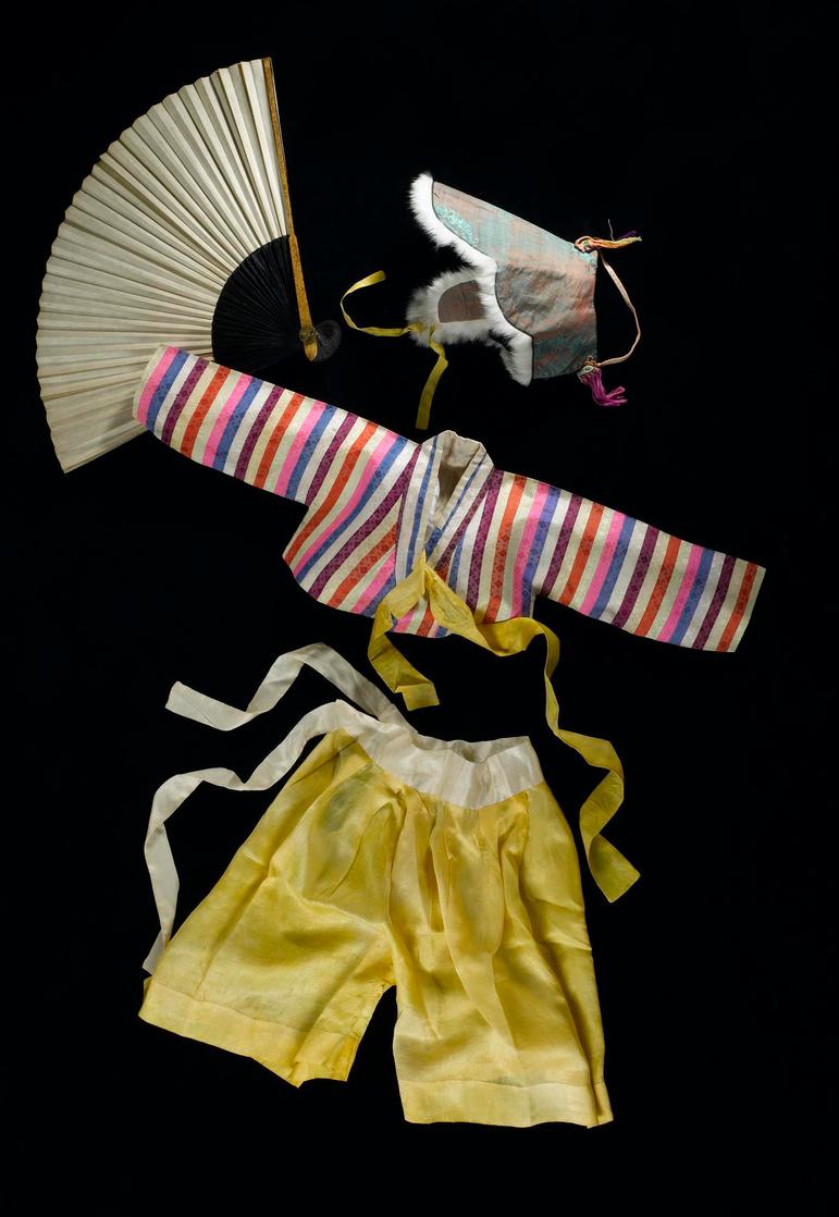 A child's outfit and accessories laid flat including a fur lined hat, a cropped stripy bolero jacket, yellow wide trousers, and a folding paper fan.
