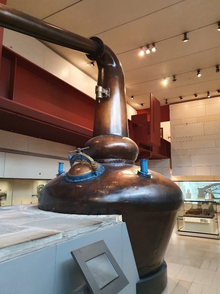 A large, round, copper vessel, wide at the bottom and with a pipe protruding from the top that curves 90 degrees further up. The still is in a modern gallery space.