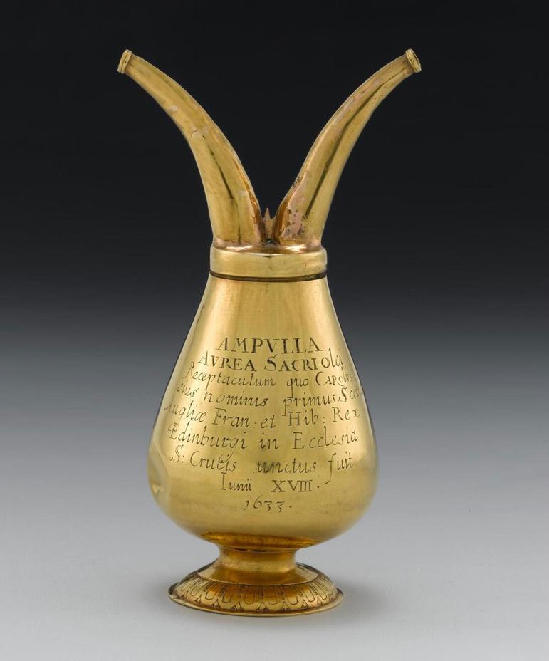A short-stemmed gold vessel, wide at the bottom and narrow top with two hoses attached to the top and engraved text to the body of the vessel.