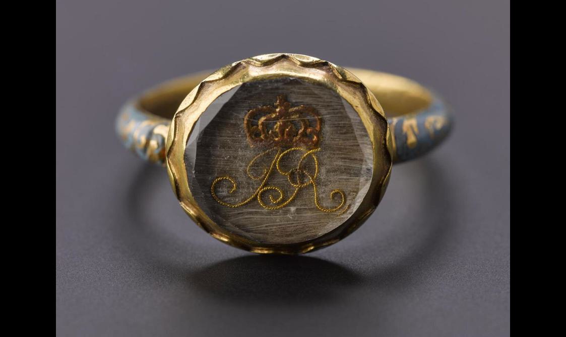 A close shot of an enamel ring showing the circle inset within a glass with a monogram "JR" and a crown above the initials. The outer edge of the glass is decorated with geometric band made out of gold.