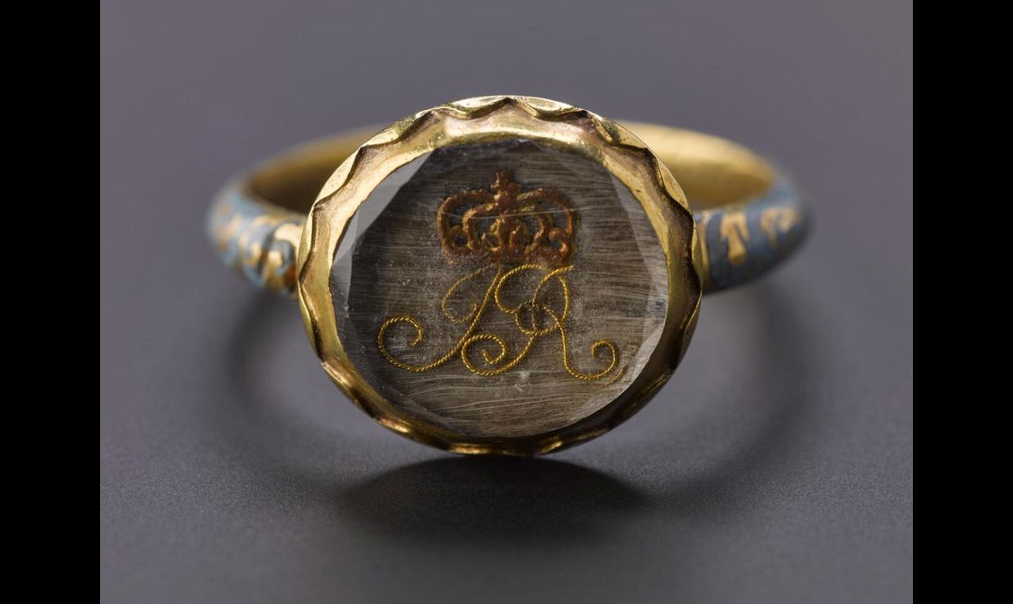 A close shot of an enamel ring showing the circle inset within a glass with a monogram "JR" and a crown above the initials. The outer edge of the glass is decorated with geometric band made out of gold.