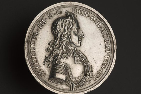 A silver coin with inscription to the edge and a profile portrait of a man in shoulder length curly hair wearing metal armour.