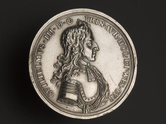 A silver coin with inscription to the edge and a profile portrait of a man in shoulder length curly hair wearing metal armour.