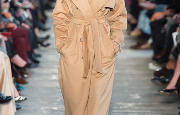 A model wearing a beige hijab, long coat, and brown trousers on a catwalk runway.