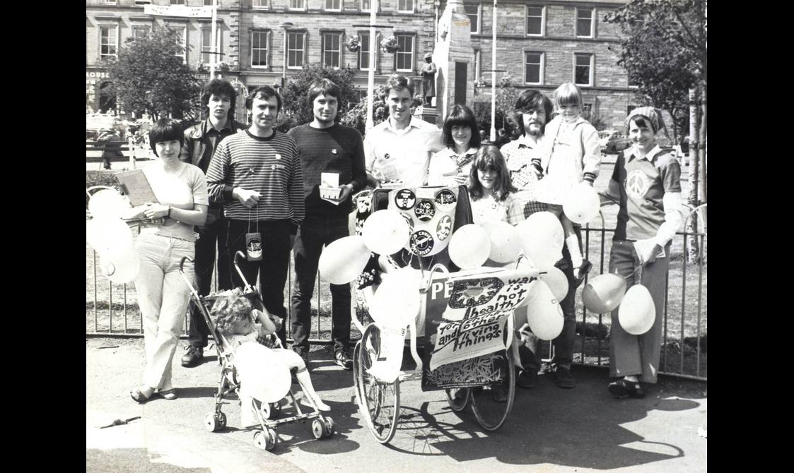 A black and white photo of a group of people standing in a city square holding balloons. There is a child's pram covered in protest signs and buttons.
